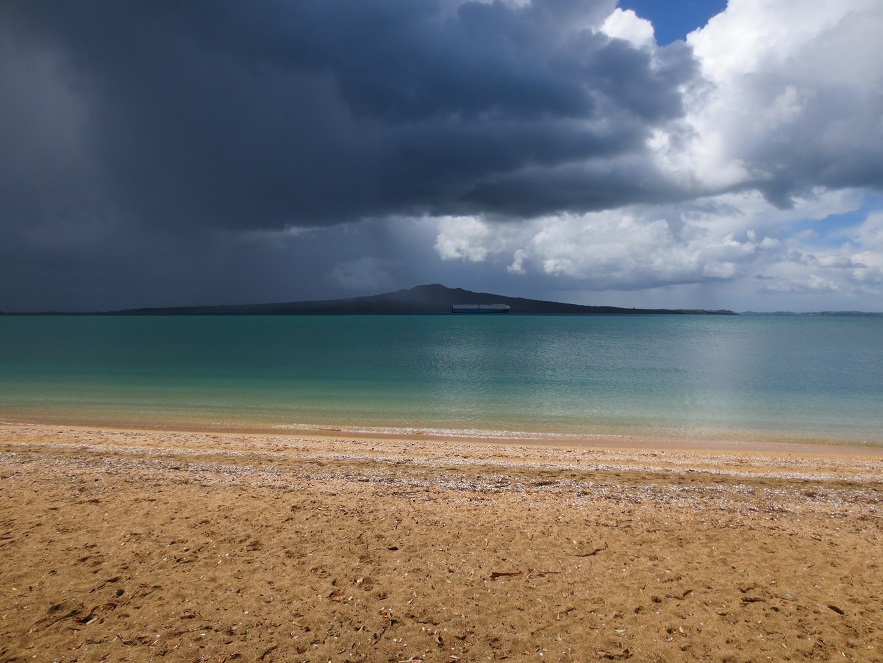 Volcanic cone island and storm clouds on horizon; blue-green sea in mid-ground; beach in foreground.