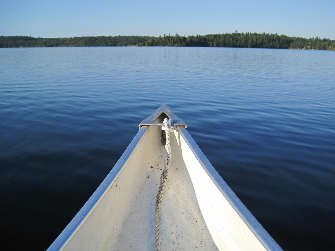 Canoe prow in foreground bisecting an expanse of blue lake water, framed by distant spruce trees