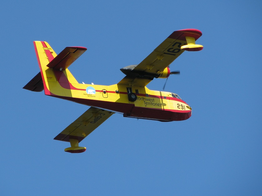 Airborne NWT water bomber in yellow and red colours against bright blue sky